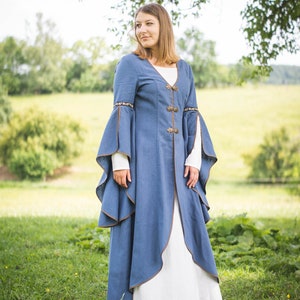 Op de afbeelding: Een vrouw die een blauwe middeleeuwse jurk draagt met bruine bies en decoratieve knopen. De jurk heeft lange mouwen met een vloeiende vorm. Ze staat in een grasveld.