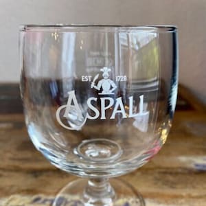 Könnte beinhalten: Klares Bierglas mit Stiel und weißem Schriftzug "ASPALL". Das Glas hat eine runde Schale und einen kurzen Stiel. Der Text "EST 1728" befindet sich über einer Grafik eines Kochs. Das Glas steht auf einer Holzoberfläche.