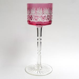 May include: A tall, elegant wine glass with a vibrant pink bowl and a clear, intricately cut stem and base. The bowl features a detailed, etched design. The glass is designed for serving wine.