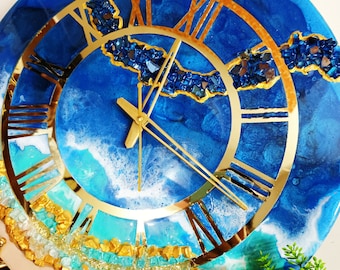 May include: A round wall clock featuring a blue and white resin pour background that mimics ocean waves. The clock face has gold-colored Roman numerals and hands, accented with embedded dark blue and gold crushed glass textures.