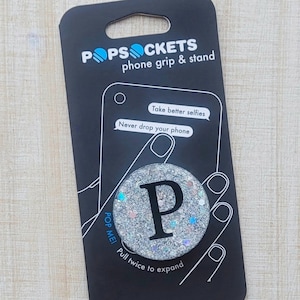 May include: A black phone grip and stand packaging with the brand name "POPSOCKETS" printed on it. The packaging shows a silver glitter phone grip with a black letter "P" in the center. The text "Take better selfies" and "Never drop your phone" is printed on the packaging.