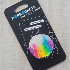 May include: A PopSockets phone grip and stand with a rainbow glitter design. The black packaging features the PopSockets logo and the text "Take better selfies" and "Never drop your phone". The grip is white, with a rainbow gradient and glitter.