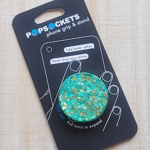 May include: A turquoise and gold glitter phone grip and stand with the text "POPSOCKETS phone grip & stand" and "Take better selfies. Never drop your phone. POP ME! Pull twice to expand."