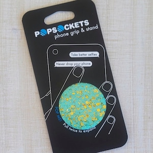 May include: A turquoise blue PopSocket with gold glitter. The PopSocket is on a black phone grip and stand packaging with the text "POPSOCKETS phone grip & stand" and "Take better selfies Never drop your phone POP ME! Pull twice to expand".