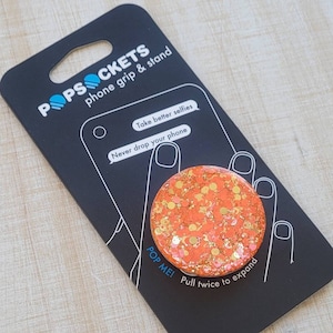 May include: A black phone grip and stand with the text "POPSOCKETS phone grip & stand" and "Take better selfies Never drop your phone POP ME! Pull twice to expand". The stand is orange with gold glitter.