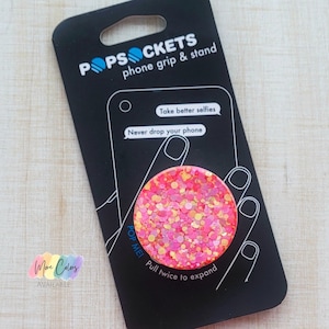 May include: A pink and gold glitter PopSocket phone grip and stand. The packaging features the text "POPSOCKETS phone grip & stand", "Take better selfies", "Never drop your phone", "POP ME! Pull twice to expand", and "More Colors AVAILABLE".