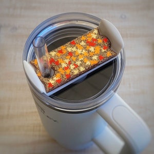 May include: A white travel mug with a clear lid and a straw. The lid features a rectangular accent piece decorated with autumn leaves in shades of red, orange, and gold. The mug has a white handle.