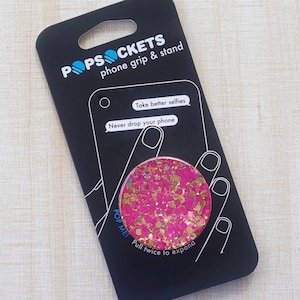 May include: Pink and gold glitter phone grip and stand with the text "POPSOCKETS phone grip & stand", "Take better selfies", "Never drop your phone", "POP MEI", and "Pull twice to expand".