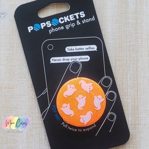 May include: A black Popsocket phone grip and stand with a Halloween theme. The orange circular grip features pink ghost designs. The packaging includes the text "Take better selfies" and "Never drop your phone."