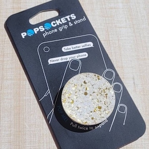 May include: A white and gold PopSocket phone grip and stand. The PopSocket has a white background with gold glitter and the words "Pop Mel" printed on the side. The packaging says "Take better selfies" and "Never drop your phone".