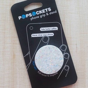 May include: A white PopSockets phone grip with a glittery finish. The packaging features the text "POPSOCKETS phone grip & stand", "Take better selfies", "Never drop your phone", and "POP MEI Pull twice to expand".