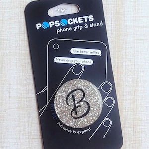 May include: A silver glitter phone grip and stand with the letter "B" in a black script font. The packaging says "PopSockets phone grip & stand" and "Pull twice to expand".