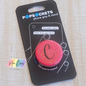 May include: A pink glitter phone grip and stand with the letter "C" in white. The packaging says "PopSockets phone grip & stand" and "Pop Me! Pull twice to expand".