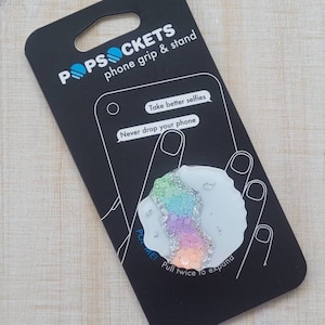 May include: A white PopSockets phone grip with a rainbow glitter design. The packaging says "PopSockets phone grip & stand", "Take better selfies", "Never drop your phone", and "Pull twice to expand".
