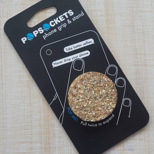 May include: A gold glitter PopSocket phone grip and stand. The packaging features the text "POPSOCKETS" and "phone grip & stand". It also says "Take better selfies" and "Never drop your phone". The PopSocket says "POP ME!" and "Pull twice to expand".