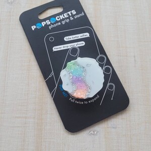 May include: A white PopSockets phone grip and stand with a rainbow glitter design. The packaging says "Take better selfies" and "Never drop your phone".
