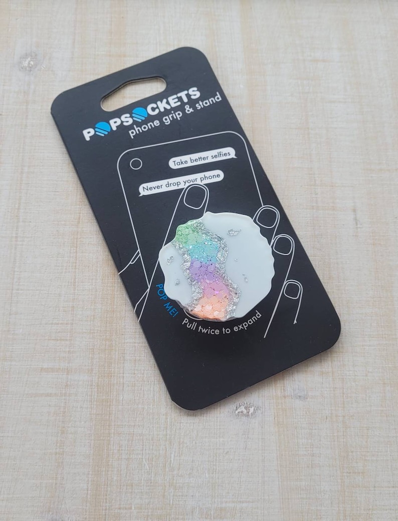 May include: A white PopSockets phone grip and stand with a rainbow glitter design. The packaging says "Take better selfies" and "Never drop your phone".