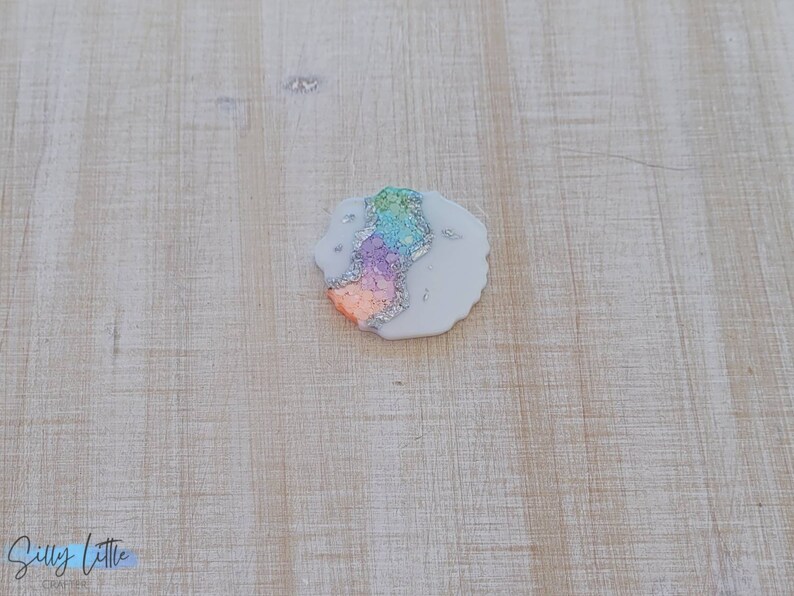 May include: A white resin coaster with a colorful agate design. The coaster has a rainbow of colors including pink, orange, yellow, green, and blue. The coaster is covered in silver glitter.