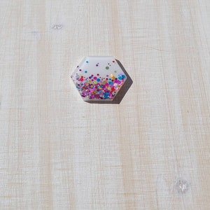 以下が含まれることがあります： カラフルなグリッターが埋め込まれた六角形の透明な樹脂製のペーパーウェイトです。