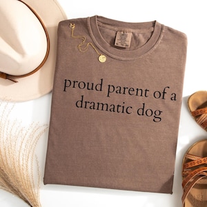 Puede incluir: Una camiseta marrón con el texto "proud parent of a dramatic dog" en negro. Un collar dorado y un sombrero color crema están junto a la camiseta. En la imagen también hay sandalias con correas marrones.