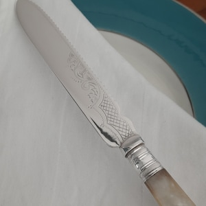 May include: A silver-colored serrated knife with ornate etched designs on the blade. The handle is a light, pearlescent color. The knife is resting on a white cloth, with a teal-rimmed plate in the background.
