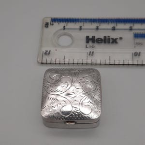 May include: A small, silver, rectangular box with a hinged lid. The box is decorated with a floral pattern. A clear ruler with centimetre markings is in the background.