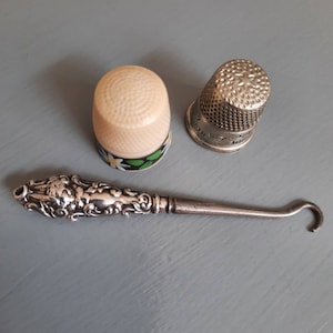 May include: A collection of sewing implements, featuring two thimbles and a silver crochet hook. One thimble is cream-coloured with a floral pattern, the other is silver. The crochet hook has a decorative handle.