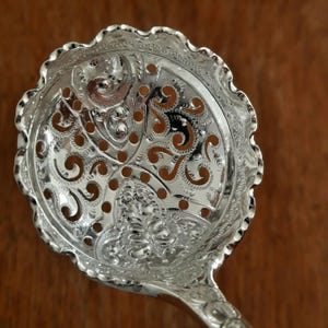 May include: Ornate silver serving spoon with a decorative, pierced bowl. The bowl features intricate scrollwork and a scalloped edge. The handle is also detailed with similar patterns. The spoon is designed for serving and adds elegance to any table setting.
