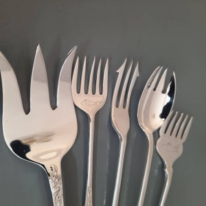 May include: A set of five silver-toned metal serving forks of varying shapes and sizes. The largest fork has three wide tines, while the others feature intricate designs including a fish engraving, pointed tips, and a rounded spoon-like shape.