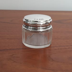 May include: A small, round glass jar with a clear, vertically ribbed body and a polished silver lid. The lid features a series of small, stamped hallmarks visible on its top surface. The container has a wide, flat base and a smooth, reflective finish.
