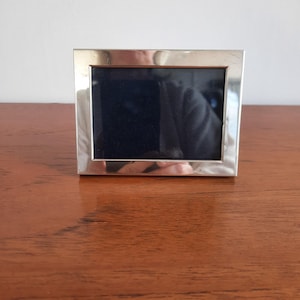 May include: A rectangular picture frame with a polished silver-tone metallic finish and a wide border. The frame features a dark, empty centre display area, reflecting the surrounding environment on its smooth, reflective surface.