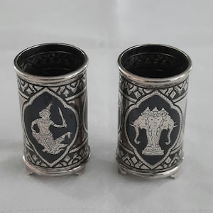 May include: Two cylindrical metal cups featuring black enamel panels with intricate silver-toned engravings. One cup displays a figure holding a sword, while the other depicts a three-headed elephant. Both vessels stand on small, raised feet.
