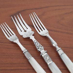May include: Three silver forks with white mother-of-pearl handles. Each fork features a unique metal design at the base of the tines, including floral and scrollwork engravings. The handles are smooth and rectangular with rounded edges.