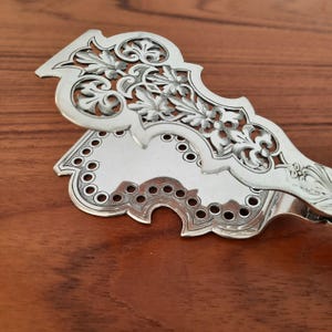 May include: A silver-toned metal serving tong featuring an intricate openwork floral pattern on the top handle and a scalloped edge with a series of small circular cutouts along the perimeter of the lower gripping surface.