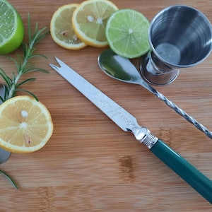 May include: A stainless steel bar knife with a green handle, a double-sided jigger, a spoon, and a long mixing spoon are arranged on a wooden cutting board. Sliced lemons and limes, and sprigs of rosemary are also present.