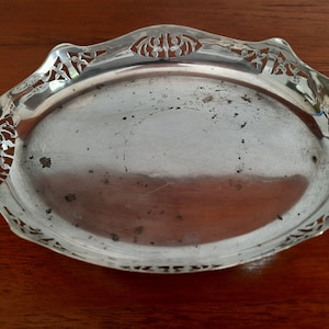 May include: Oval silver-toned serving tray with decorative cut-out edges. The tray has a slightly tarnished surface and is set against a dark wooden background. The edges feature intricate floral and geometric patterns.