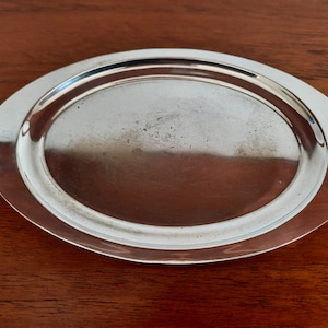 May include: A shiny, oval-shaped silver serving tray. The tray has a slightly raised rim and a smooth, reflective surface. The tray is set against a dark brown wooden surface.