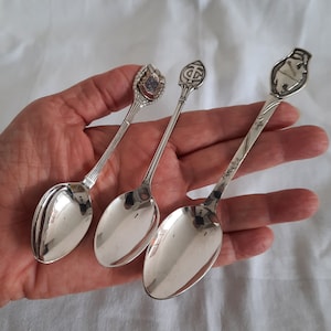 May include: Three silver-toned metal souvenir spoons with decorative handles. The left spoon features a colourful crest, the centre spoon has a shield-shaped top with the letters TC, and the right spoon displays a raised figure of a bear.