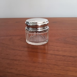 May include: A small, clear glass jar with a faceted, multi-sided base and a polished silver-tone metal lid. The lid features a smooth, rounded top surface with small, engraved hallmarks visible near the edge of the metallic cap.