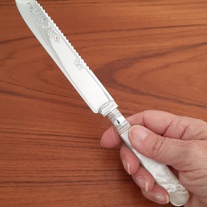 May include: A silver butter knife with a serrated edge and ornate floral engraving on the blade. The handle is white and has a pearlescent finish. The knife is held in a hand.