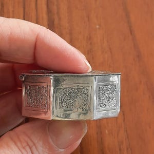 May include: A small, hexagonal silver-colored metal box featuring a hinged lid and detailed, embossed rectangular panels on each side. The panels display a textured, intricate pattern of foliage and branches, with a polished metallic finish.