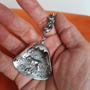 May include: A silver-toned metal scoop featuring a handle topped with a small figure of a winged creature. The wide, triangular bowl of the scoop displays a relief design of a cow standing in a field with trees and a fence in the background.