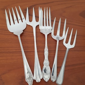 May include: A collection of five silver-toned metal forks with varying designs. The set includes one wide four-tined fork, two three-tined forks with ornate handles, one three-tined fork with a leaf engraving, and one slender three-tined fork.
