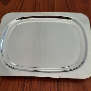 May include: A rectangular, silver-colored serving tray with rounded corners. The tray has a smooth, reflective surface and a slightly raised edge. The tray is empty and sits on a wooden surface.