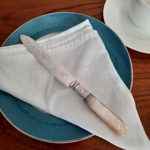 May include: A silver butter knife with an ornate floral design on the blade rests on a white napkin atop a teal plate. The handle is a light cream colour. A white teacup and saucer are in the background.