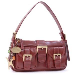 May include: A dark red leather shoulder bag featuring two front pockets with brass-toned buckles, a central buckle closure, and a top handle. A dangling charm set includes a gold-toned cat figure and a metal tag embossed with the text CATWALK.
