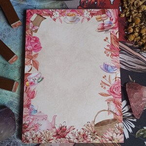 May include: A blank notepad with a floral border featuring teacups, teapots, butterflies, and macarons. The border is in shades of pink, red, and green.