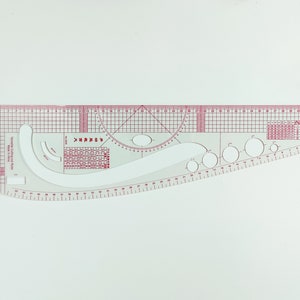 May include: A clear, pink, plastic sewing ruler with various curved and straight edges, along with a grid and circular cutouts. The ruler has measurements in centimetres, and Chinese text.