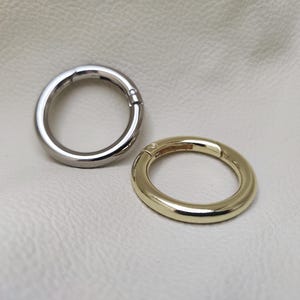 Könnte beinhalten: Zwei Metallringe, einer silberfarben und einer golden, mit einem Drehverschluss. Die Ringe liegen auf einem weißen Hintergrund.