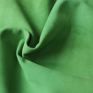 Può includere: Primo piano di un tessuto morbido di colore verde con una texture liscia. Il tessuto è piegato e drappeggiato, creando un motivo fluido e ondulato.